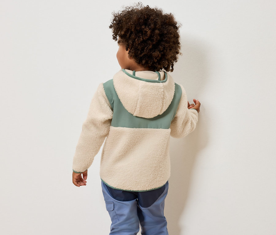 Rückenansicht eines Kindes mit lockigem Haar, das eine cremefarbene Fleecejacke mit Kapuze und grünen Akzenten sowie blaue Hosen trägt.