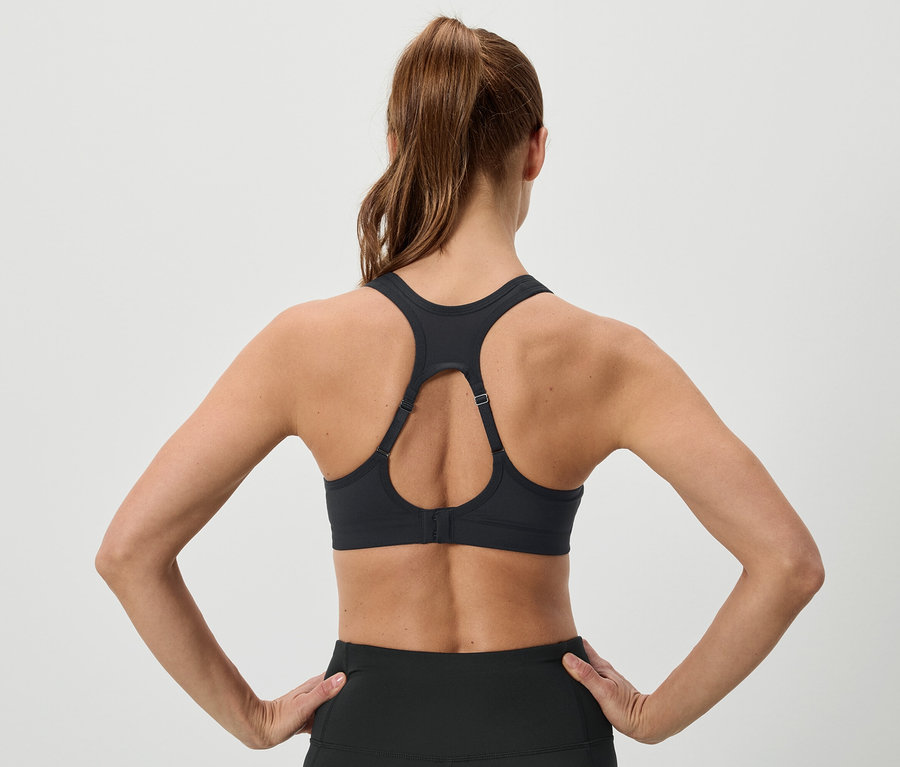 Rückenansicht einer Frau mit hochgesteckten braunen Haaren, die einen schwarzen Sport-BH und Leggings trägt und die Hände in die Hüften stemmt.