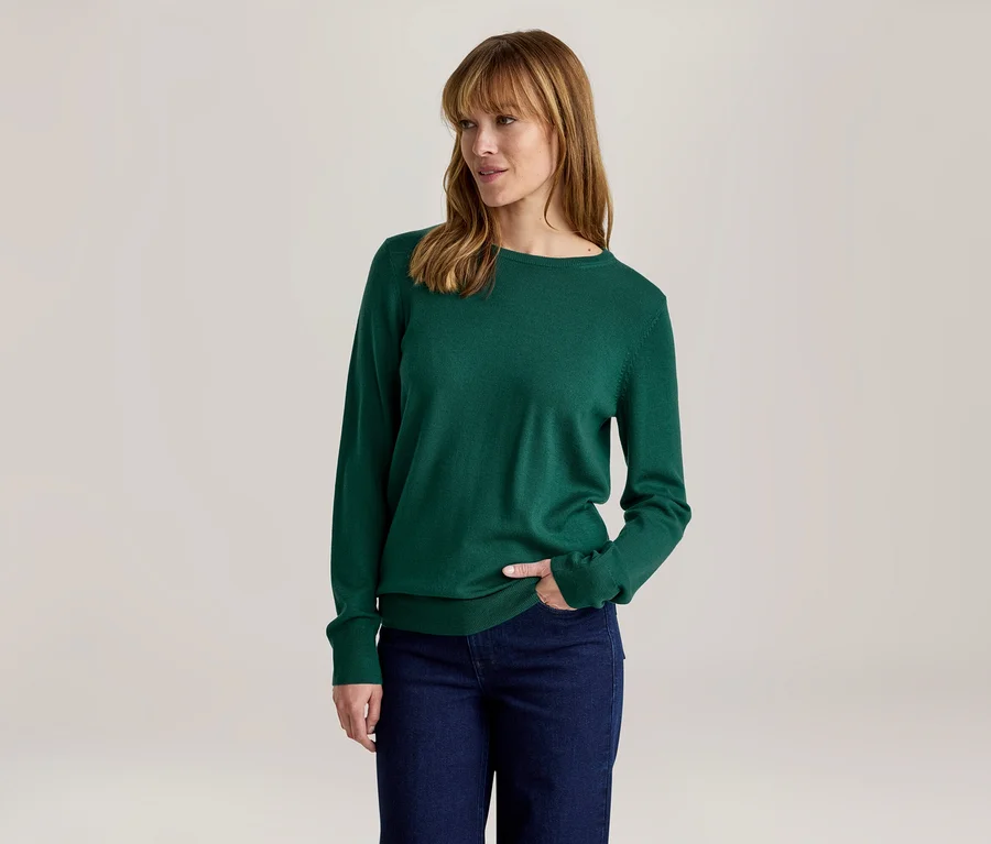 Frau in einem grünen Langarmshirt und dunklen Jeans, mit der rechten Hand in der Tasche, blickt nach rechts.