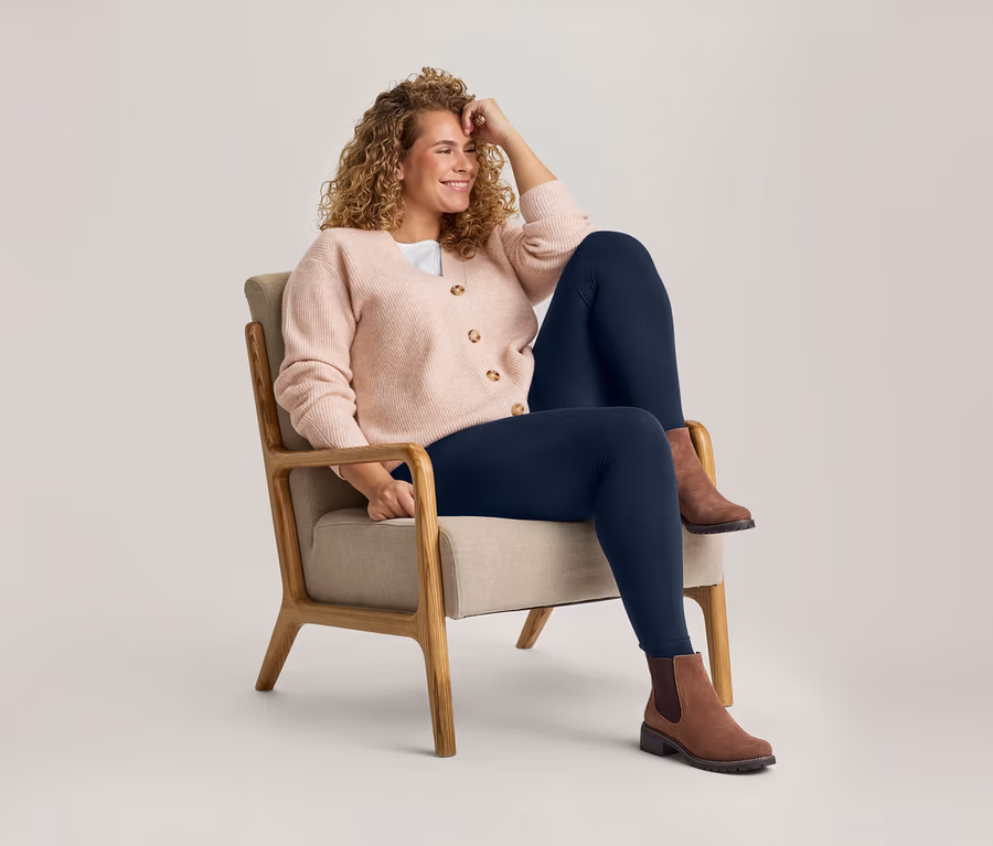Eine lächelnde Frau mit lockigem Haar sitzt in einem Sessel, trägt eine hellrosa Strickjacke, ein weißes Oberteil, dunkelblaue Leggings und braune Stiefeletten.