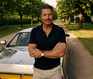 Mann mit verschränkten Armen vor einem Auto, trägt ein schwarzes Poloshirt, beigefarbene Hose und eine goldfarbene Kienzle Uhr »Meister Klassik« auf einer von Bäumen gesäumten Straße.