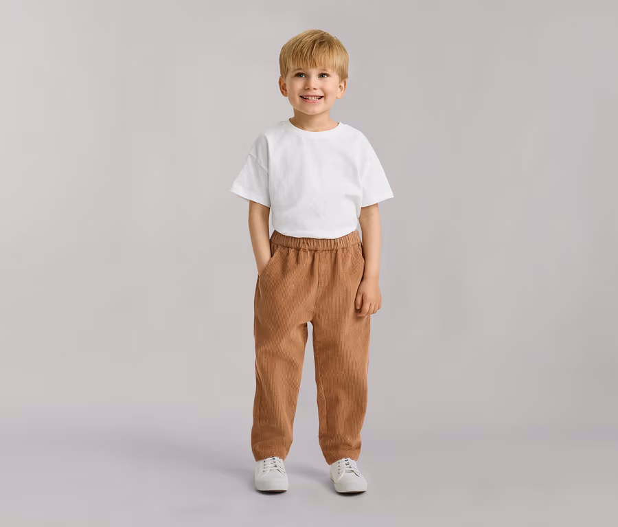 Junger Junge mit blondem Haar, lächelnd, trägt ein weißes T-Shirt, braune Cordhose und weiße Turnschuhe, Hände in den Taschen.