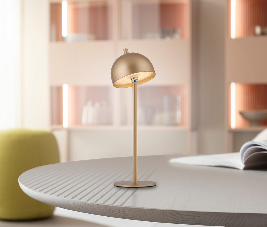 Goldfarbene SCHÖNER WOHNEN Akku-Tischleuchte »Bellis« mit Kuppelschirm auf einem hellen, geriffelten Holztisch, im Hintergrund ein verschwommenes Wohnzimmer.