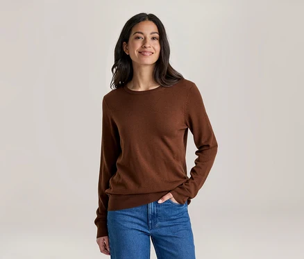 Eine lächelnde Frau mit dunklen Haaren trägt ein braunes Langarmshirt und blaue Jeans, eine Hand in der Tasche, vor hellem Hintergrund.