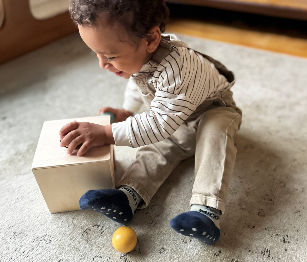 Kleinkind sitzt auf einem Teppich und spielt mit dem small foot Montessori-Bundle 3, einem Holzkasten und einem gelben Ball. Das Kind trägt ein gestreiftes Shirt und eine Latzhose und greift in den Kasten.