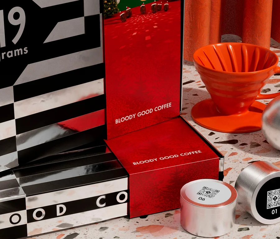Eine Nahaufnahme von Kaffeezubehör: Eine schwarz-silberne Box mit „19 grams“, eine rote Box mit „BLOODY GOOD COFFEE“, ein orangefarbener Pour-Over-Kaffeefilter mit Untertasse, zwei silberne Kaffeekapseln und eine silberne Kugel auf einer gesprenkelten Oberfläche.