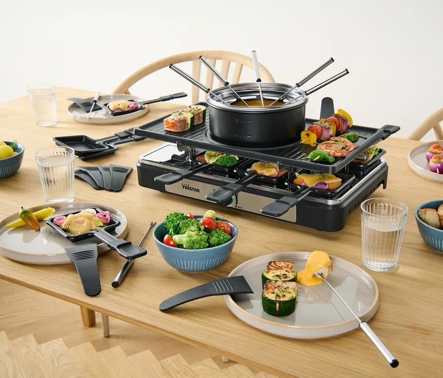 Ein 2-in-1-Raclette-und-Fondue-Gerät steht auf einem Tisch mit Speisen.