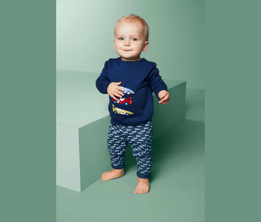 Ein Baby steht und trägt einen blauen Pullover mit Autos und eine Sweathose.