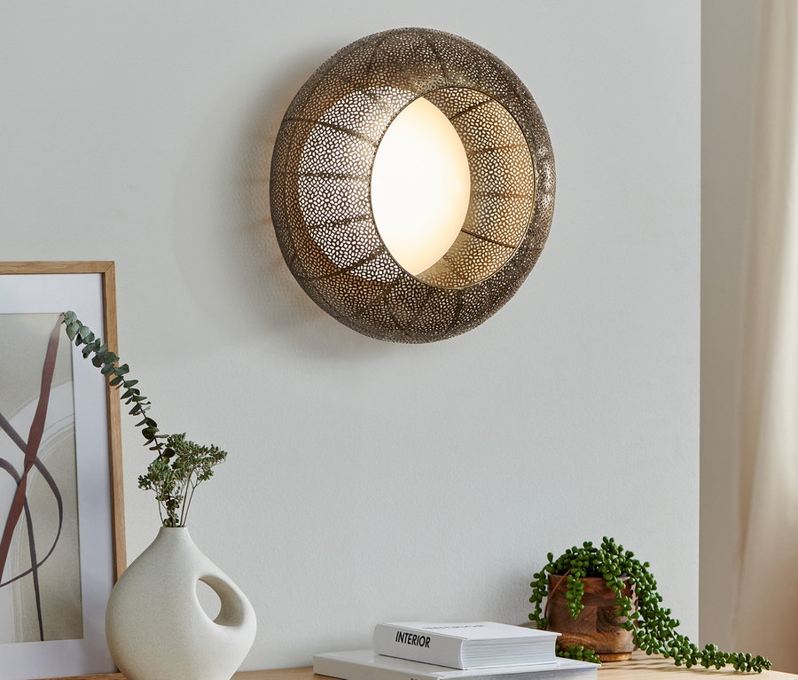 Eine LED-Wand- und Deckenleuchte hängt an der Wand. Darunter befinden sich eine Vase, eine Pflanze und Bücher auf einer Kommode.