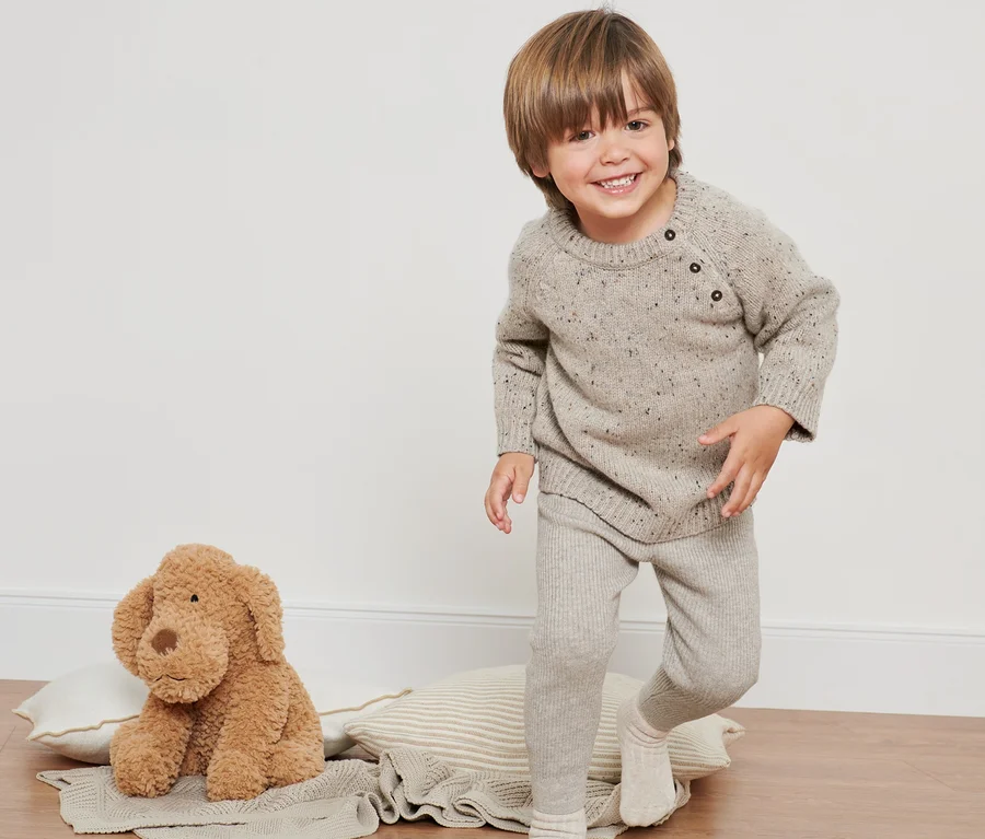 Lächelnder Junge trägt einen Strickpullover mit RWS-Wolle und Leggings. Neben ihm liegt ein Plüschhund.