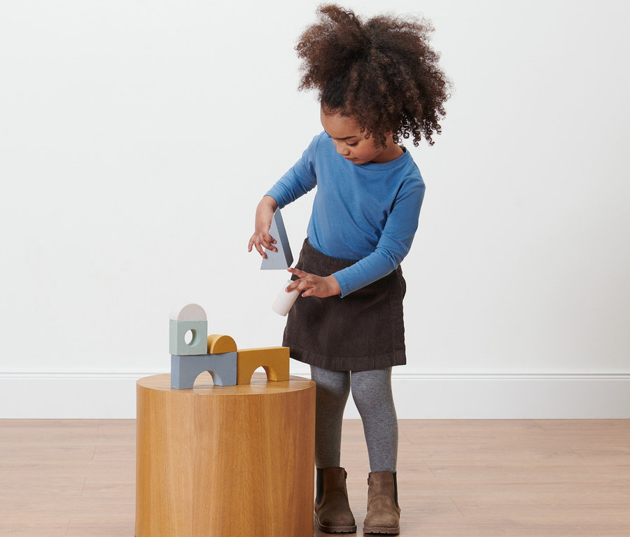 Mädchen spielt mit Holzspielzeug auf einem runden Tisch, trägt einen Kleinkind-Hosenrock.