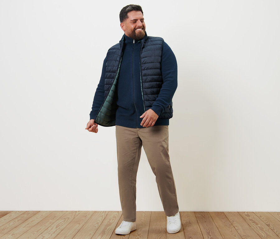 Ein lächelnder Mann steht und trägt eine Wendeweste, eine Strickjacke in Marineblau und eine sandfarbene Chino.