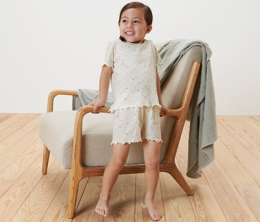 Ein lächelndes Mädchen steht an einem Sessel und trägt einen Kinder-Shorty-Pyjama.
