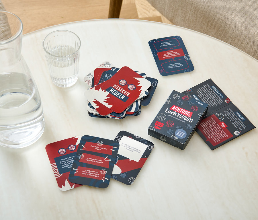 Auf einem Tisch liegen eine Glaskaraffe mit Wasser, ein Glas Wasser und Spielkarten des Kartenspiels »Achtung, Lachverbot!«.