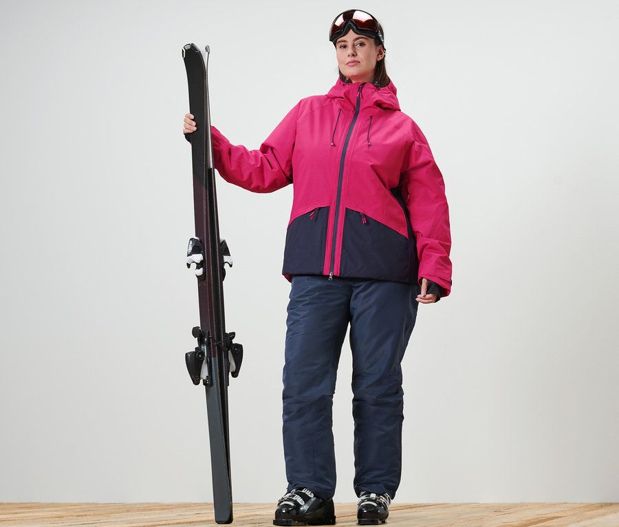 Frau hält Ski, trägt eine Skijacke und Skihose.