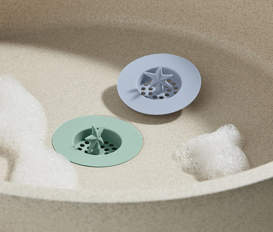 Zwei Silikon-Abflusssiebe liegen im Waschbecken, 2 Stk., eines grün und eines blau, umgeben von Schaum.