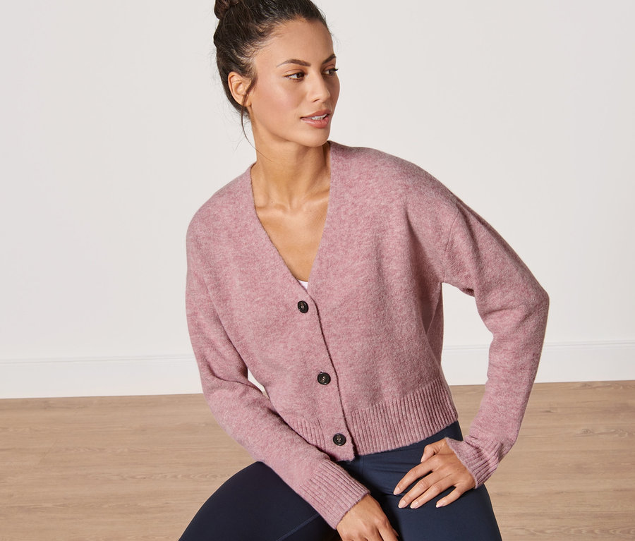 Frau sitzt auf einem Hocker und trägt einen rosa Grobstrick-Cardigan.