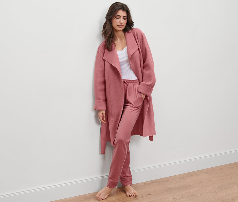 Frau lehnt an Wand und trägt Fleece-Kuschelmantel und Hose.