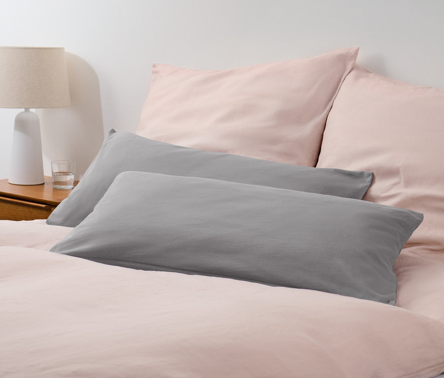 Zwei graue Jersey-Kopfkissenbezüge liegen auf einem Bett mit rosa Bettwäsche.