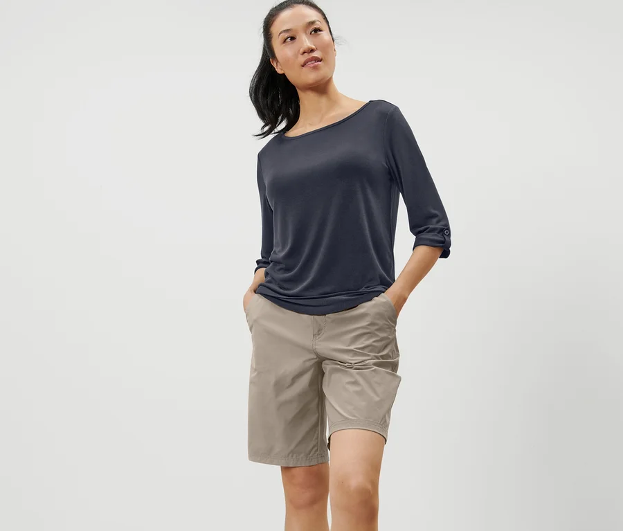 Frau steht mit Händen in den Hosentaschen und trägt ein blaues Funktionsshirt mit 3/4-Ärmeln.