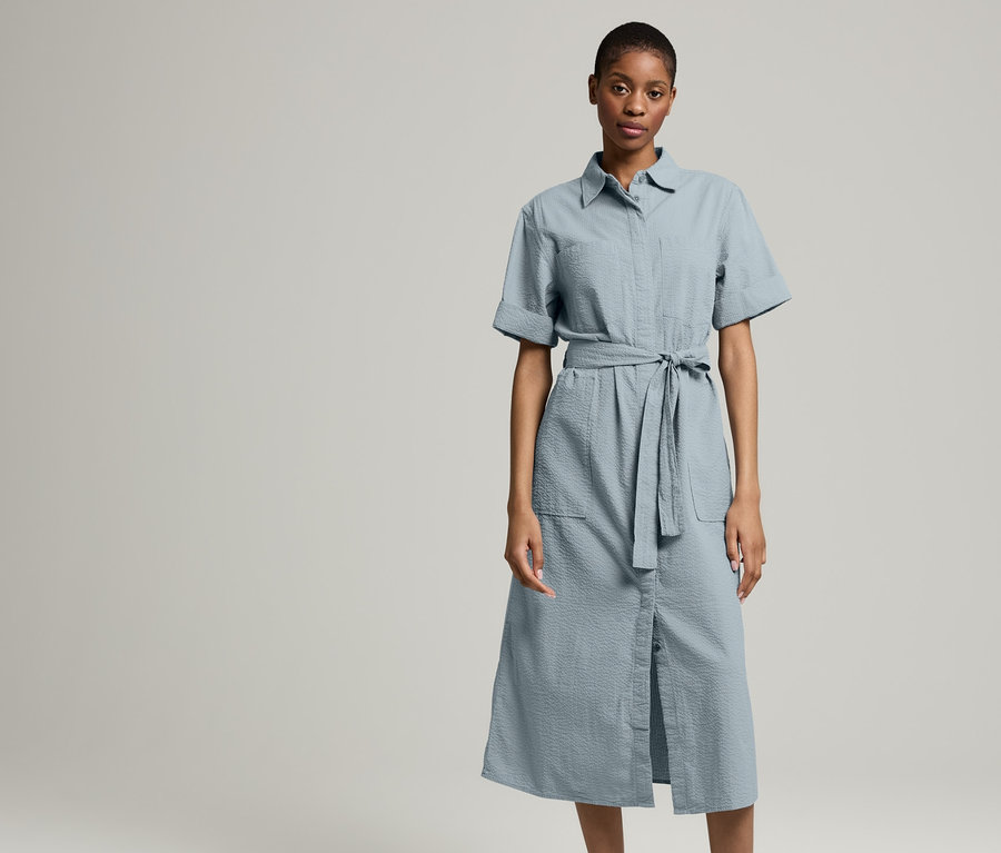Eine Frau steht vor hellem Hintergrund und trägt ein NAH/STUDIO Seersucker-Kleid | Baumwolle, Stormy Grey und eine NAH/STUDIO Jeansjacke | ungefärbte Baumwolle und Turnschuhe.