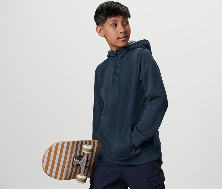 Ein Junge mit einem Skateboard trägt einen blauen Kinder-Sweat-Hoodie und schaut zur Seite.