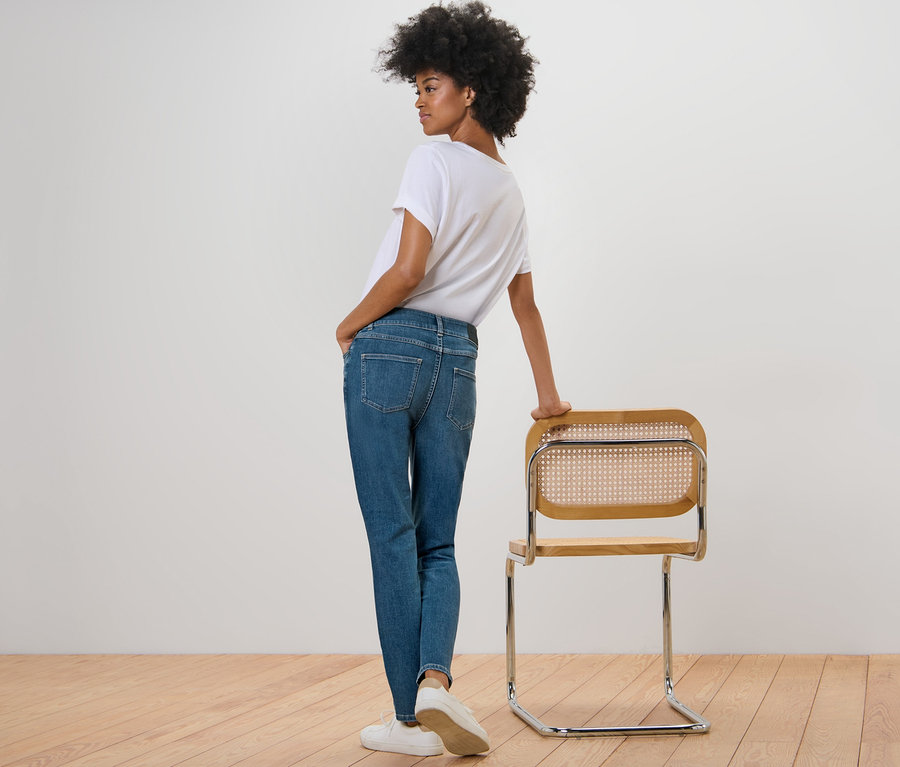 Frau posiert in Slim Jeans – Fit »Emma«, mid blue und weißem T-Shirt neben einem Stuhl.