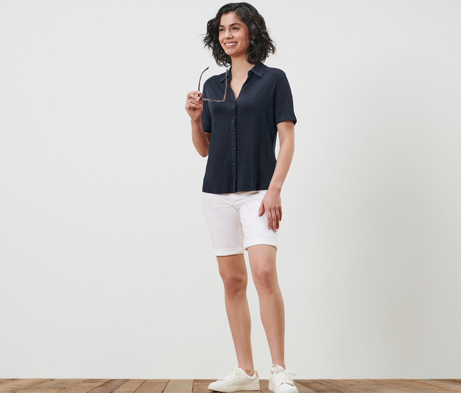 Frau hält Brille und trägt ein dunkelblaues Blusenshirt mit Knopfleiste, weiße Shorts und Turnschuhe.