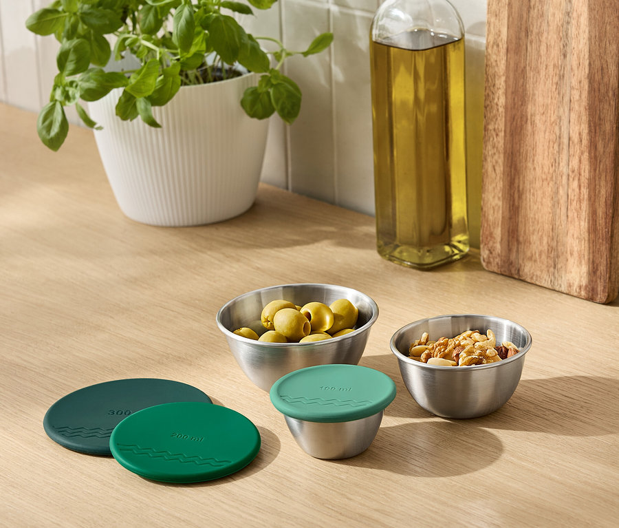 Auf einer Küchentheke stehen eine Topfpflanze, eine Ölflasche, ein Schneidebrett und Metallschalen mit Oliven und Nüssen. Eine Schale ist mit einem grünen Deckel abgedeckt.