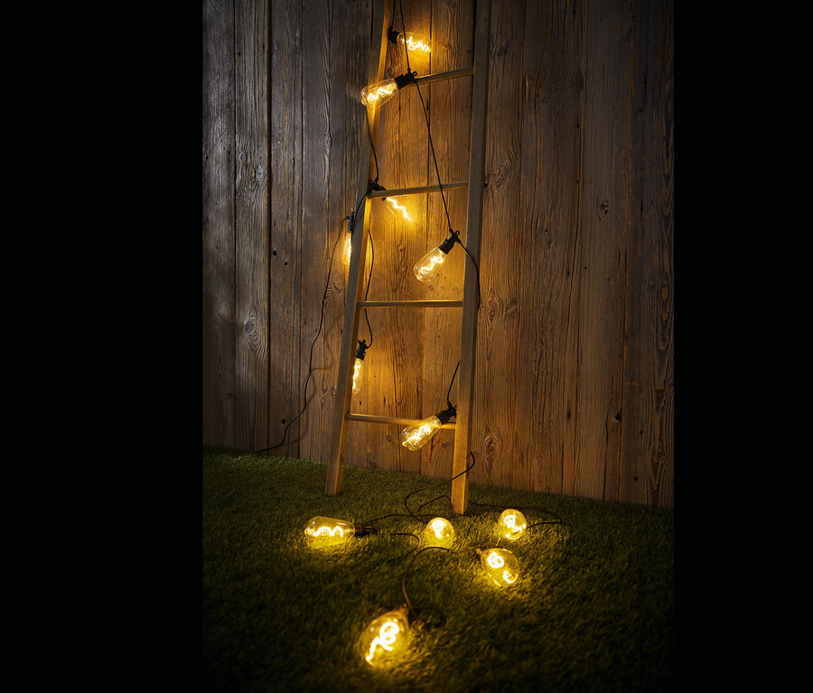 Eine Holzleiter lehnt an einer Holzwand, auf der und auf dem Boden eine LED-Filament-Lichterkette, Outdoor hängt.