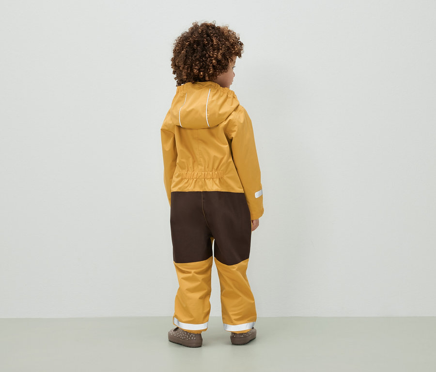 Kind mit lockigem Haar steht mit dem Rücken zur Kamera und trägt einen braun-senffarbenen Overall mit Kapuze.