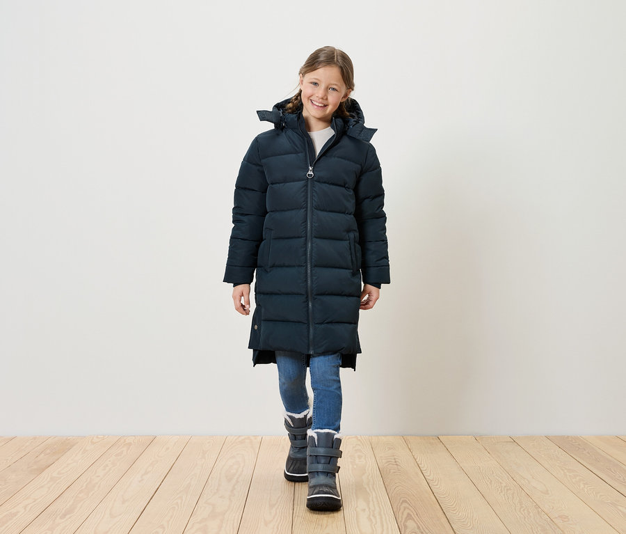 Lächelndes Mädchen in langer, gesteppter, marineblauer Jacke, blauen Jeans und grauen Winterstiefeln steht auf einem Holzboden vor einer weißen Wand.