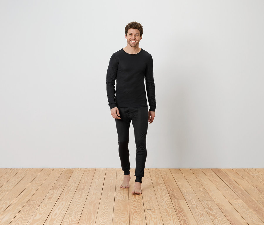 Ein Mann in schwarzer Thermounterwäsche steht barfuß auf einem Holzboden vor einer weißen Wand.