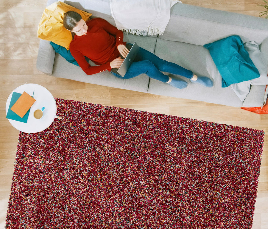 Eine Frau liegt auf einem grauen Sofa und benutzt einen Laptop. Im Vordergrund liegt ein rötlicher Teppich »Relic«, autumn spice.