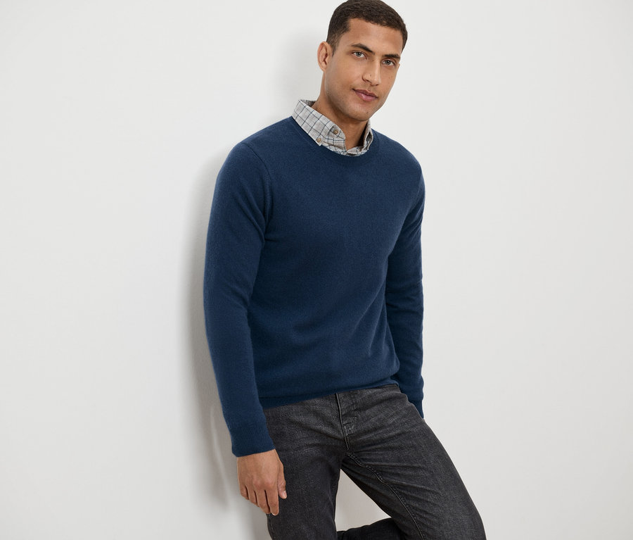 Mann lehnt an einer Wand, trägt einen blauen Pullover und eine graue gemusterte Hose.
