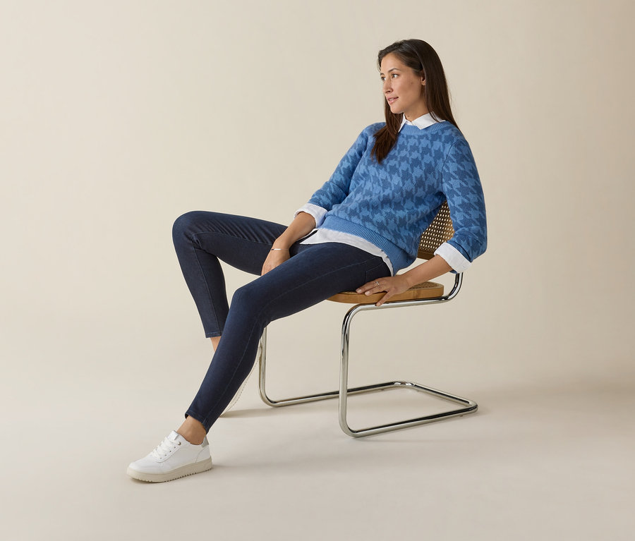 Frau sitzt auf einem Stuhl und trägt einen blauen Pullover und weiße Tom Tailor Sneaker, PU weiß.