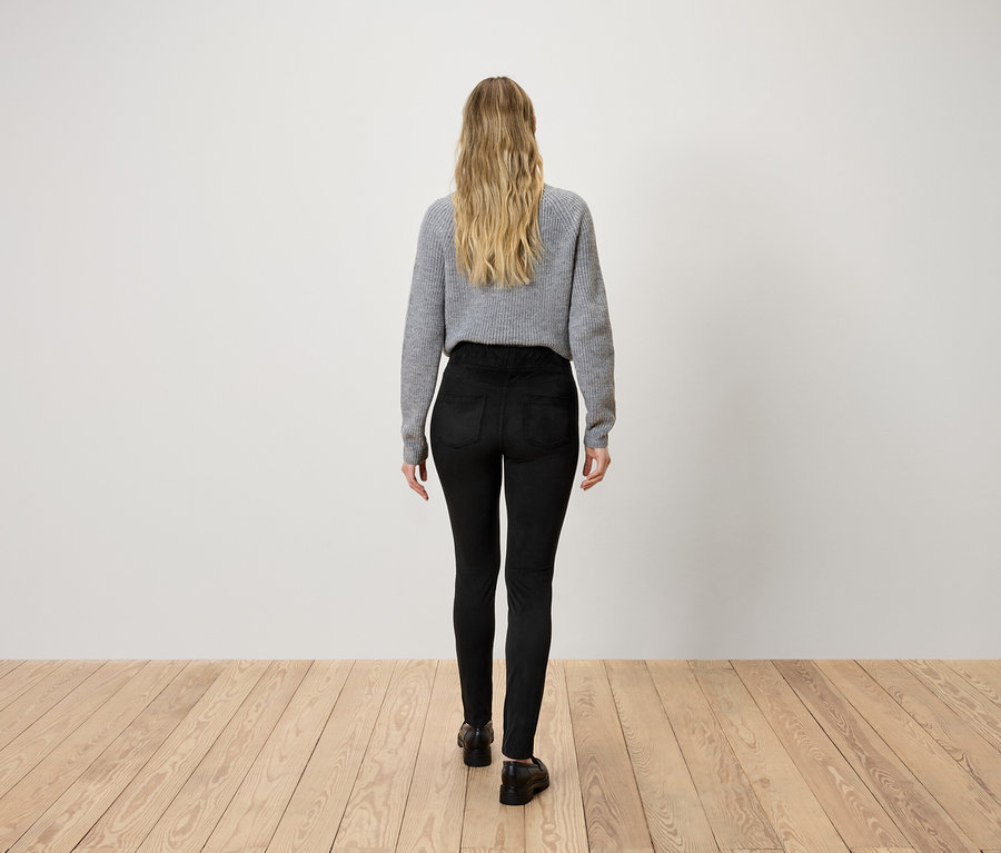 Frau mit blonden Haaren von hinten, trägt einen grauen Pullover und schwarze Hosen und geht auf einem Holzboden.