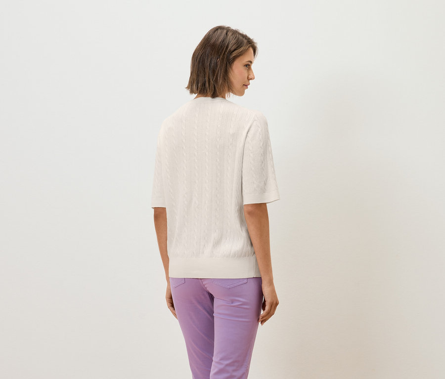 Frau mit dem Rücken zur Kamera trägt ein cremefarbenes, kurzärmeliges Strickoberteil und lila Hosen.