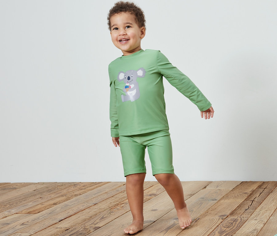 Ein Junge in Kinder-Kombi mit UV-Schutzfaktor 80, Koala, steht auf einem Holzboden neben einem Spielzeug.