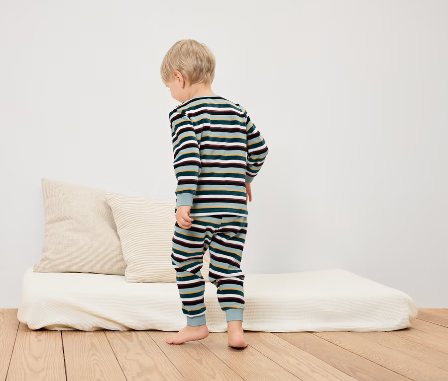 Junge steht in gestreiftem Pyjama auf dem Boden neben einem weißen Bett und Kissen.