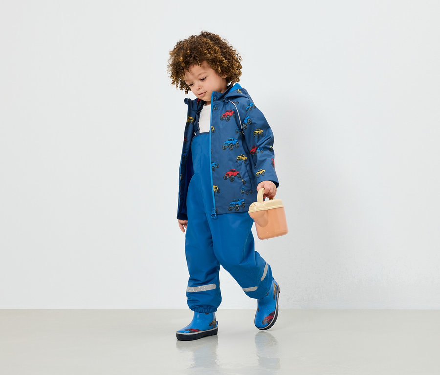 Ein Junge in blauer Latzhose, einer Jacke mit Autoaufdruck und blauen Gummistiefeln hält ein beige Spielzeug.