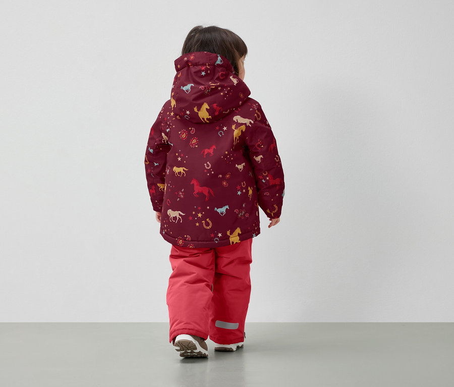Junge in Winterjacke mit Pferdeaufdruck und roten Hosen, mit dem Rücken zur Kamera.