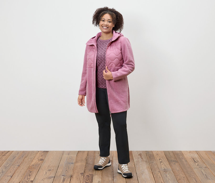 Eine Frau in einem rosafarbenen Mantel, einem gemusterten Oberteil und schwarzen Hosen steht auf einem Holzboden.