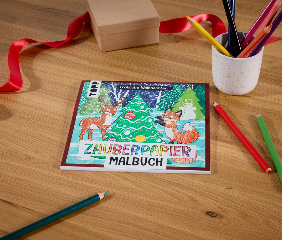 Auf einem Holztisch liegen ein Zauberpapier Malbuch mit Weihnachtsmotiv, eine Schachtel mit rotem Band, ein Becher mit Buntstiften und zwei Buntstifte.