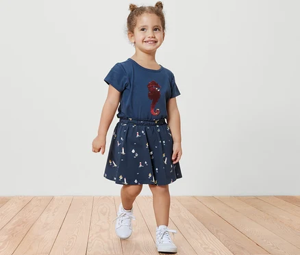 Lächelndes kleines Mädchen geht auf einem Holzboden und trägt einen Kinder-Jerseyrock mit Hose, maritim, und ein blaues T-Shirt mit einem Seepferdchen-Aufdruck.