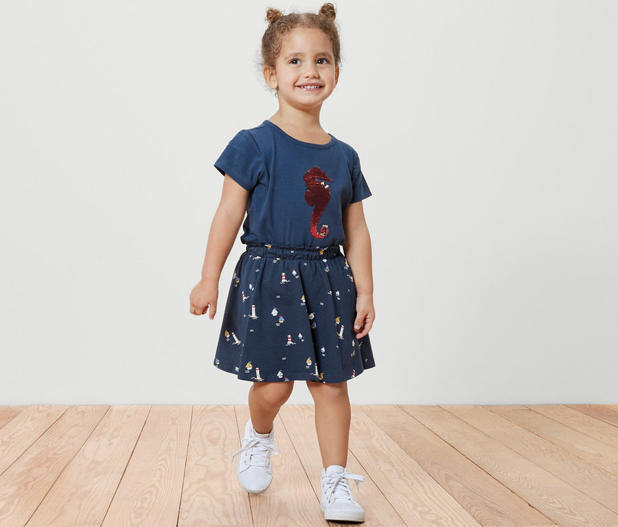 Lächelndes kleines Mädchen geht auf einem Holzboden und trägt einen Kinder-Jerseyrock mit Hose, maritim, und ein blaues T-Shirt mit einem Seepferdchen-Aufdruck.