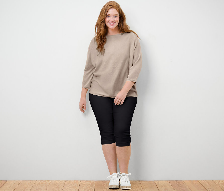 Frau steht und trägt einen beige melierten Feinstrickpullover und eine schwarze 3/4-Stretchhose.