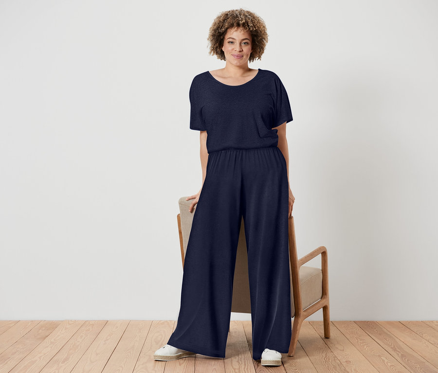 Frau mit lockigem Haar trägt einen marineblauen Jumpsuit und weiße Schuhe, während sie neben einem Stuhl mit Holzrahmen steht.