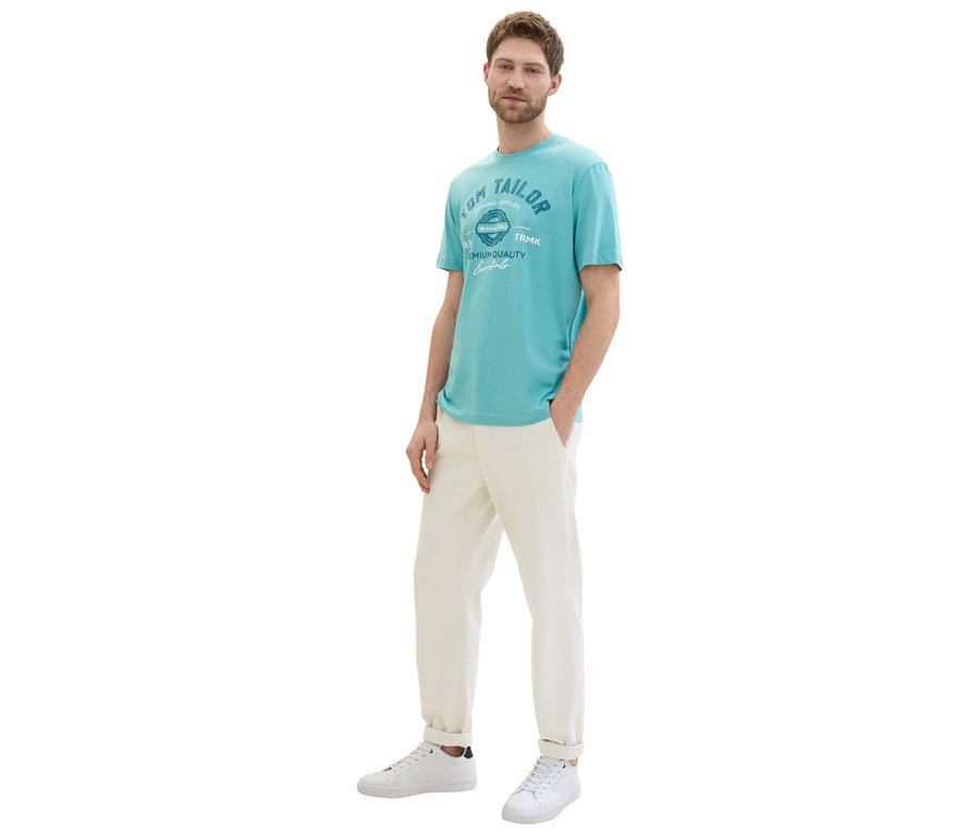 Mann in meadow teal Tom Tailor T-Shirt mit Logo, beigen Hosen und weißen Sneakers posiert.