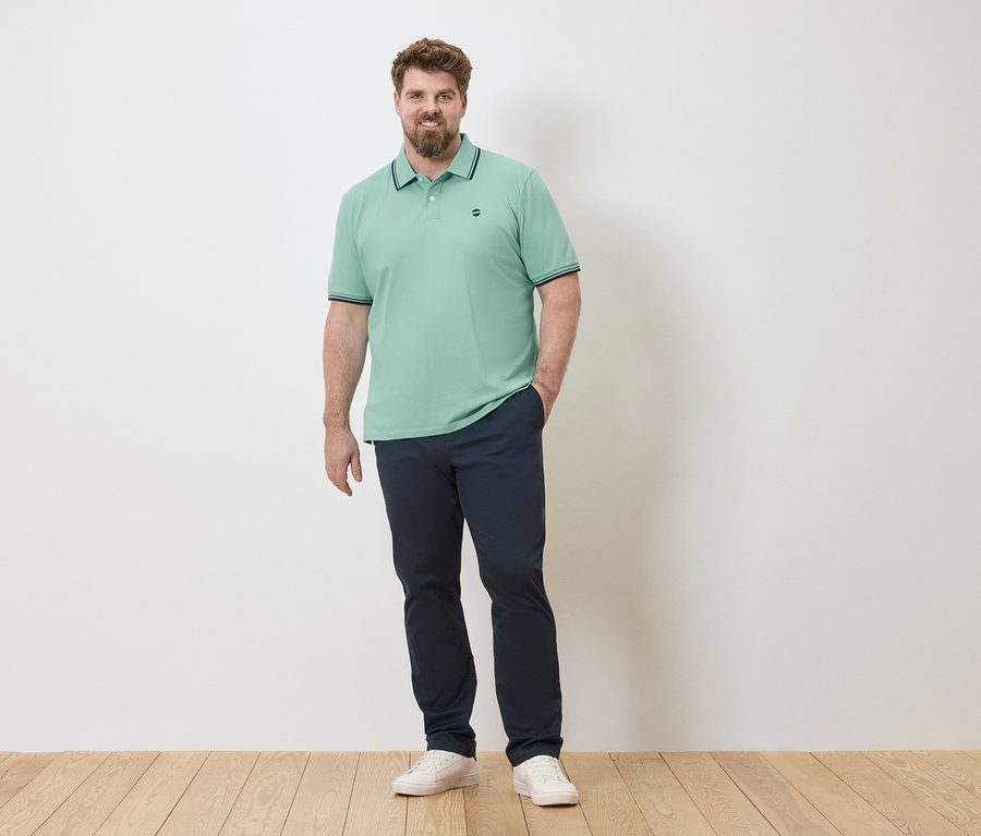 Ein Mann steht vor einer weißen Wand und trägt ein hellgrünes Poloshirt, eine marineblaue Hose und weiße Turnschuhe.